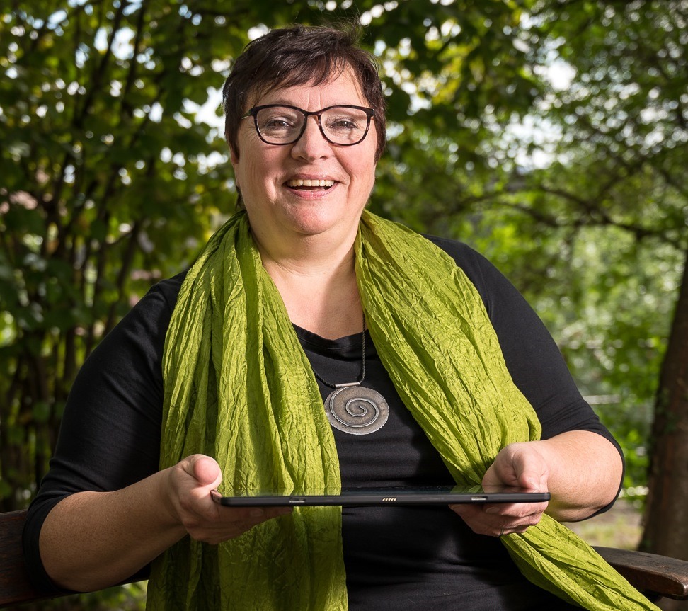 Susanne Löhnert sitzt mit einem Tablet im Grünen auf eine Parkbank