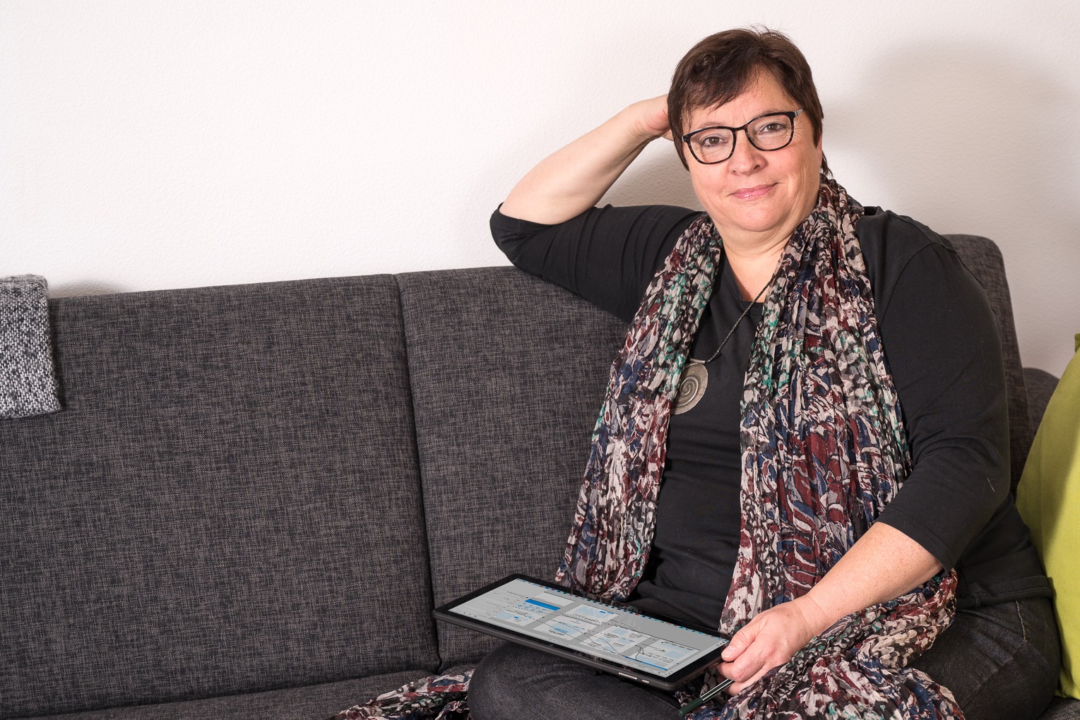 Eine Frau stitzt locker auf einer Couch mit einem Tablet auf dem Schoß und blickt offen und freundlich in die Kamera